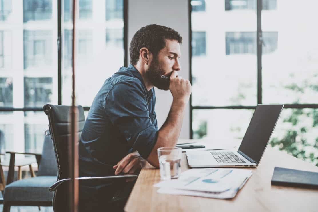 young-pensive-coworker-working-at-sunny-work-place-loft-while-sitting-at-the-wooden-table-man-analyze-document-on-laptop-display-blurred-background-horizontal