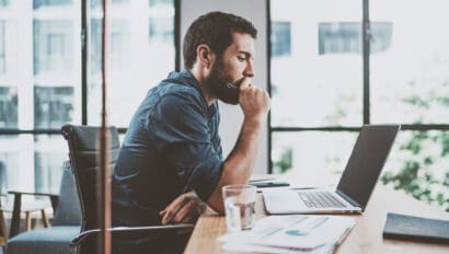 young-pensive-coworker-working-at-sunny-work-place-loft-while-sitting-at-the-wooden-table-man-analyze-document-on-laptop-display-blurred-background-horizontal